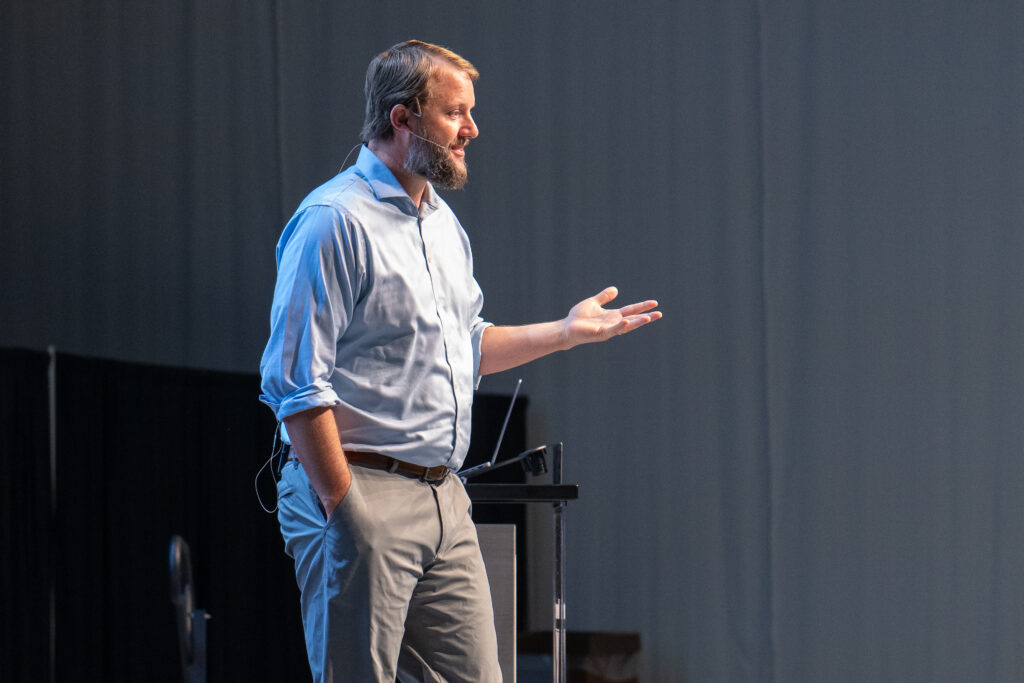 A white mal in a blue button up shirt and kahkis presents on a stage to a group of faith-based audience members.